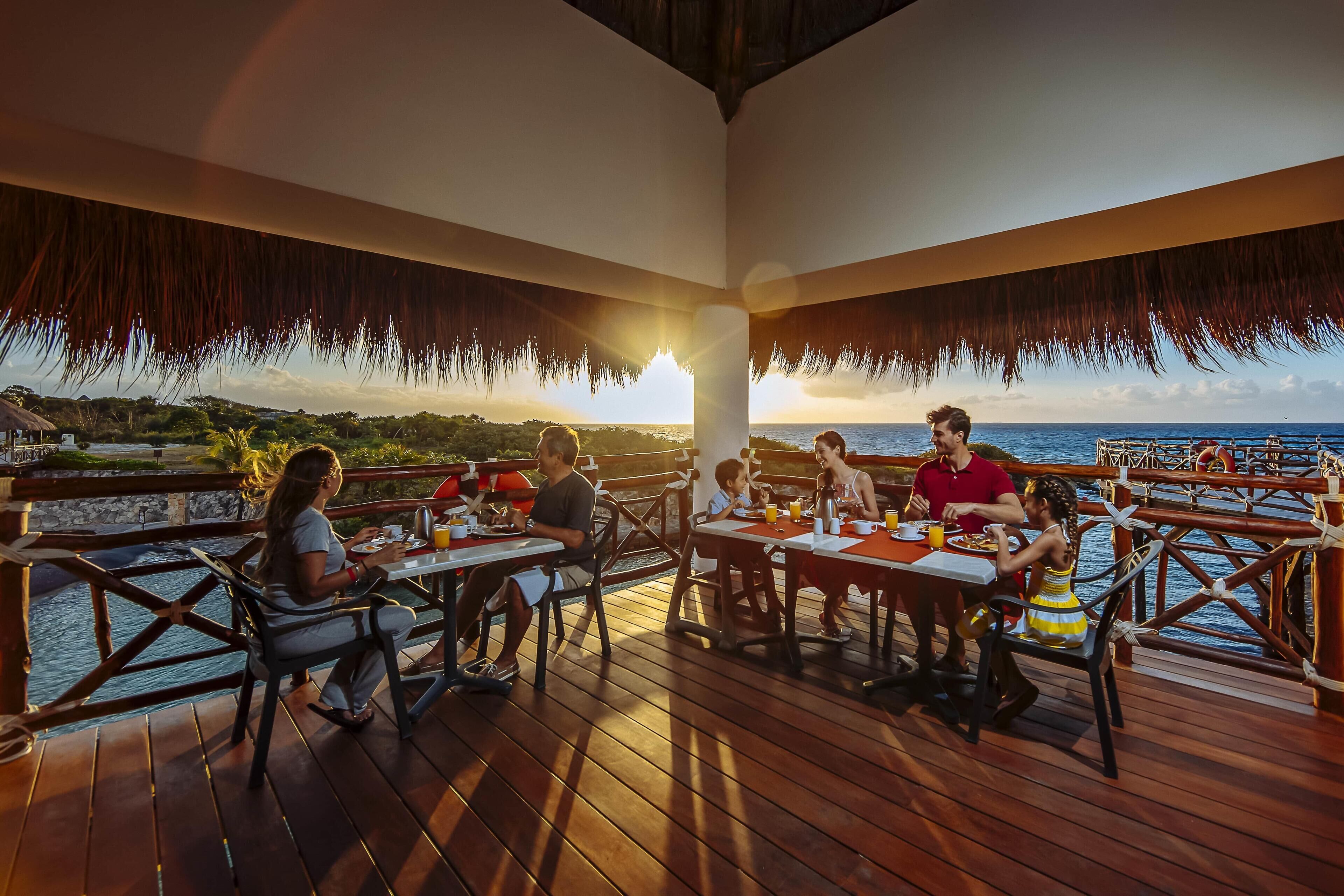 A wooden deck with tables and chairs, overlooking a body of water and a sunset.