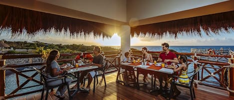 Eine Holzdielen-Terrasse mit Tischen und Stühlen, die auf ein Gewässer und einen Sonnenuntergang blickt.