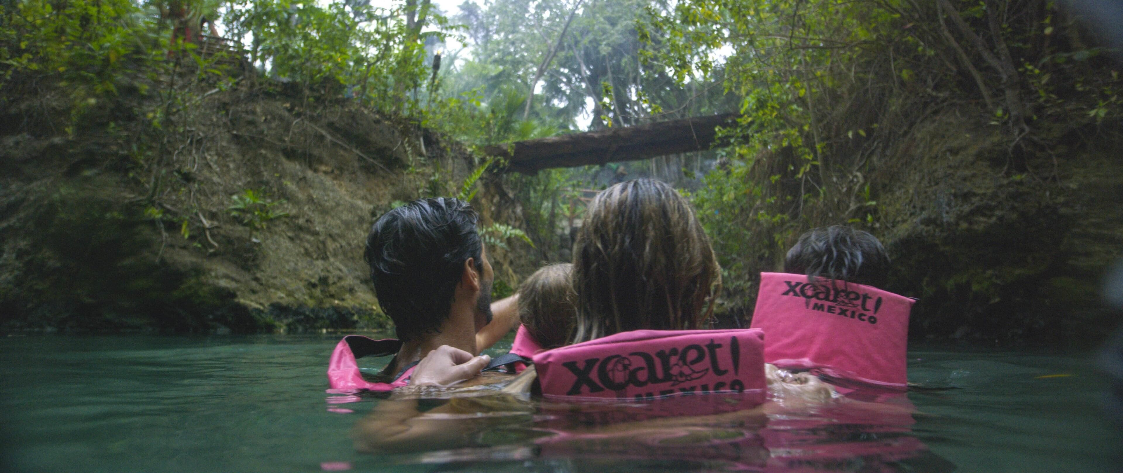 Drie mensen in een natuurlijke omgeving met water, gekleed in roze reddingsvesten met 'Xcaret'-branding.