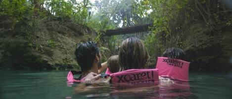 Trois personnes, dans un environnement naturel avec de l’eau, portent des gilets de sauvetage roses arborant la marque « Xcaret ».