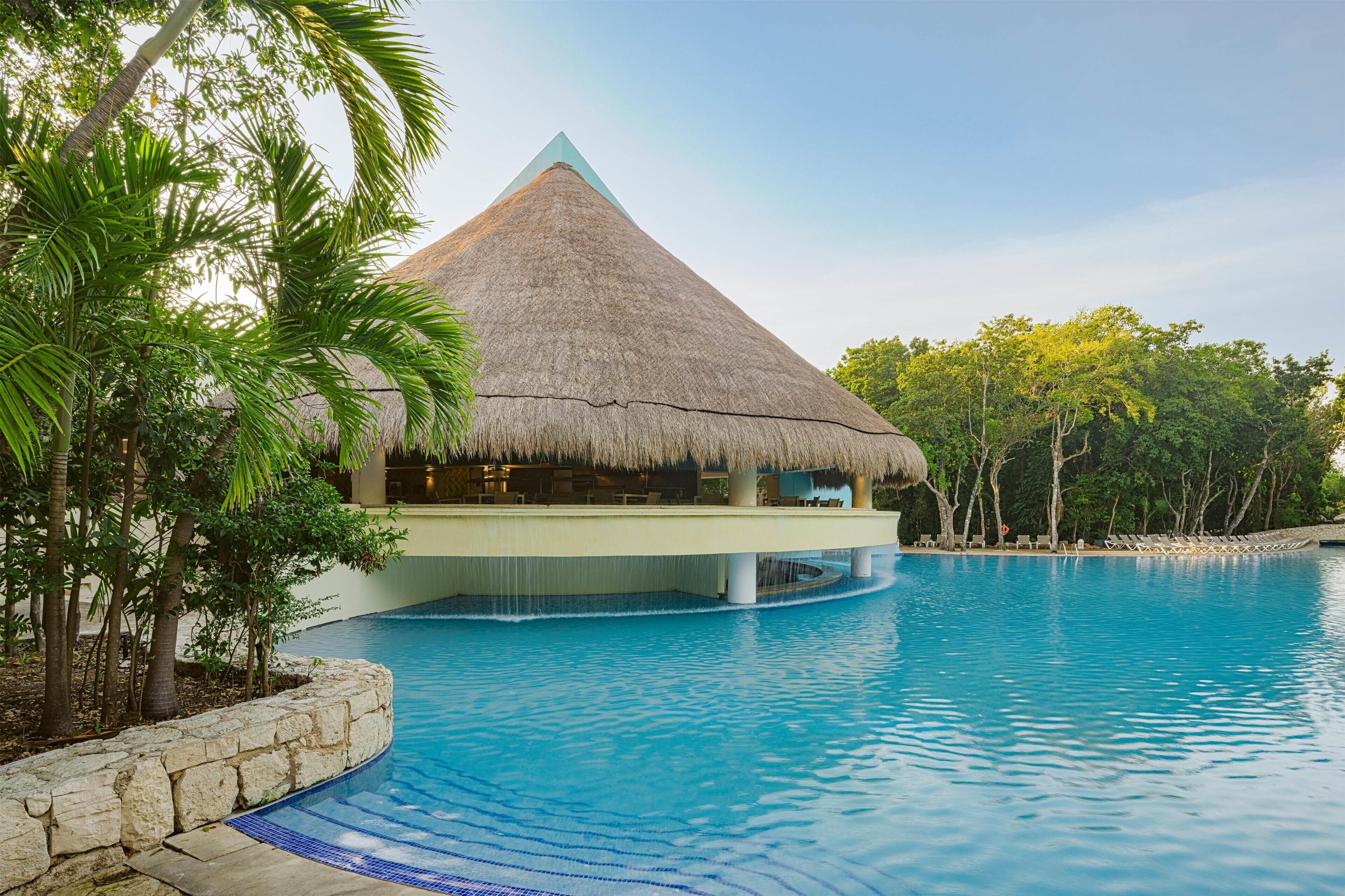A swimming pool with a thatched-roof structure, surrounded by palm trees and a stone wall.
