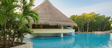 A swimming pool with a thatched-roof structure, surrounded by palm trees and a stone wall.