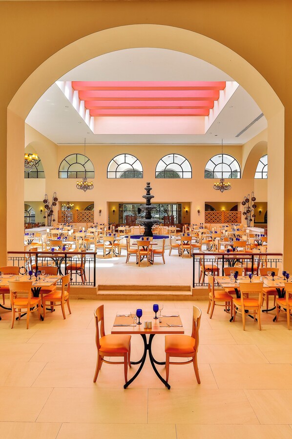 A spacious dining area with multiple tables and chairs, a central fountain, and large arched windows allowing natural light.