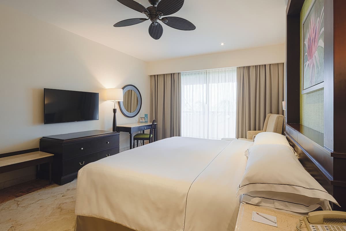 Ein Hotelzimmer mit einem großen Bett, einem Schreibtisch mit Stuhl, einem Fernseher, einem Deckenventilator und einem Fenster mit Vorhängen.