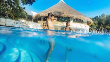Una piscina con agua cristalina azul, una estructura con techo de paja al fondo, y follaje circundante.