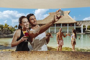 A couple taking a selfie by a pool with a thatched-roof structure in the background.