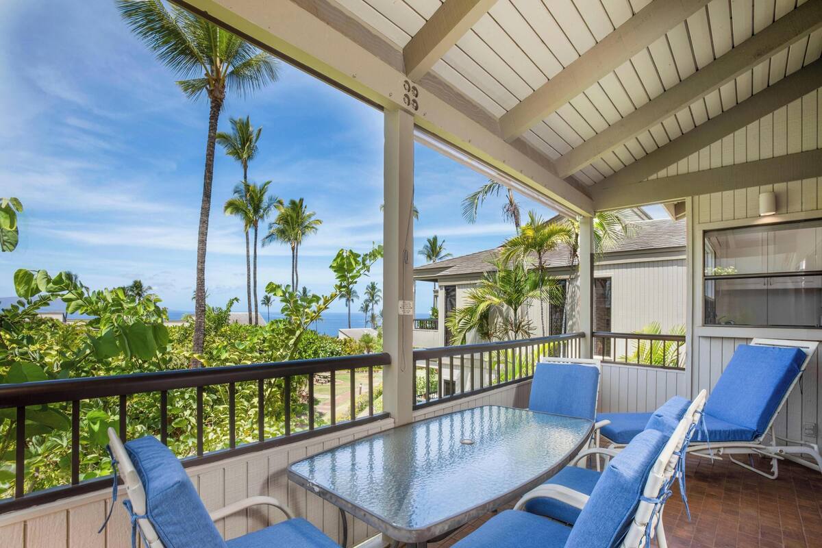 Un espace extérieur abrité, doté d’une table en verre et de chaises bleues, offrant une vue sur un paysage tropical avec des palmiers et sur l’océan.