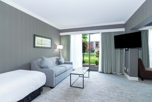 Habitación de hotel con cama, sofá, mesa de centro, televisor y vistas a un jardín a través de un ventanal corredero.
