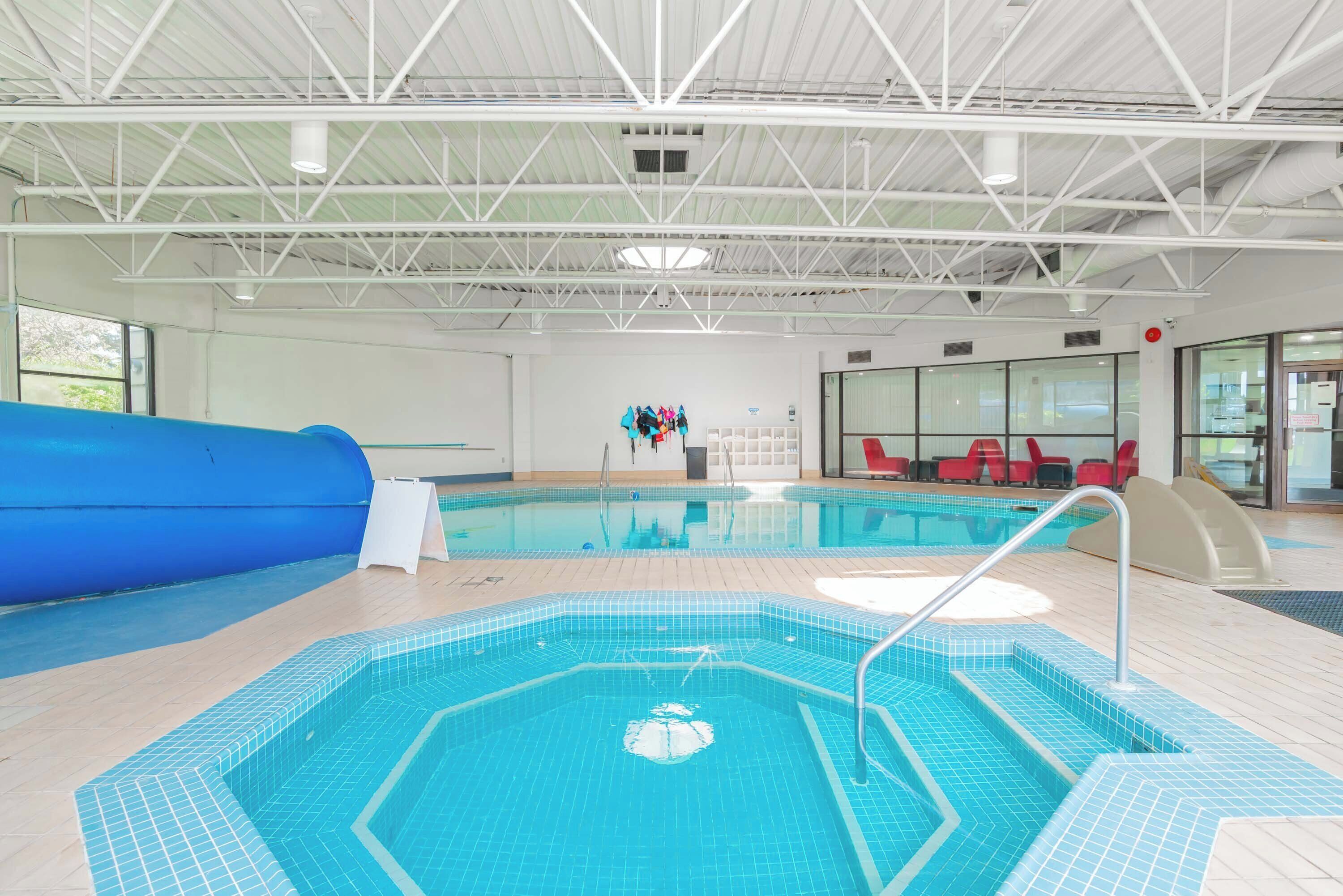 An indoor swimming pool with a blue slide, a separate hot tub area, and a seating area with red chairs.