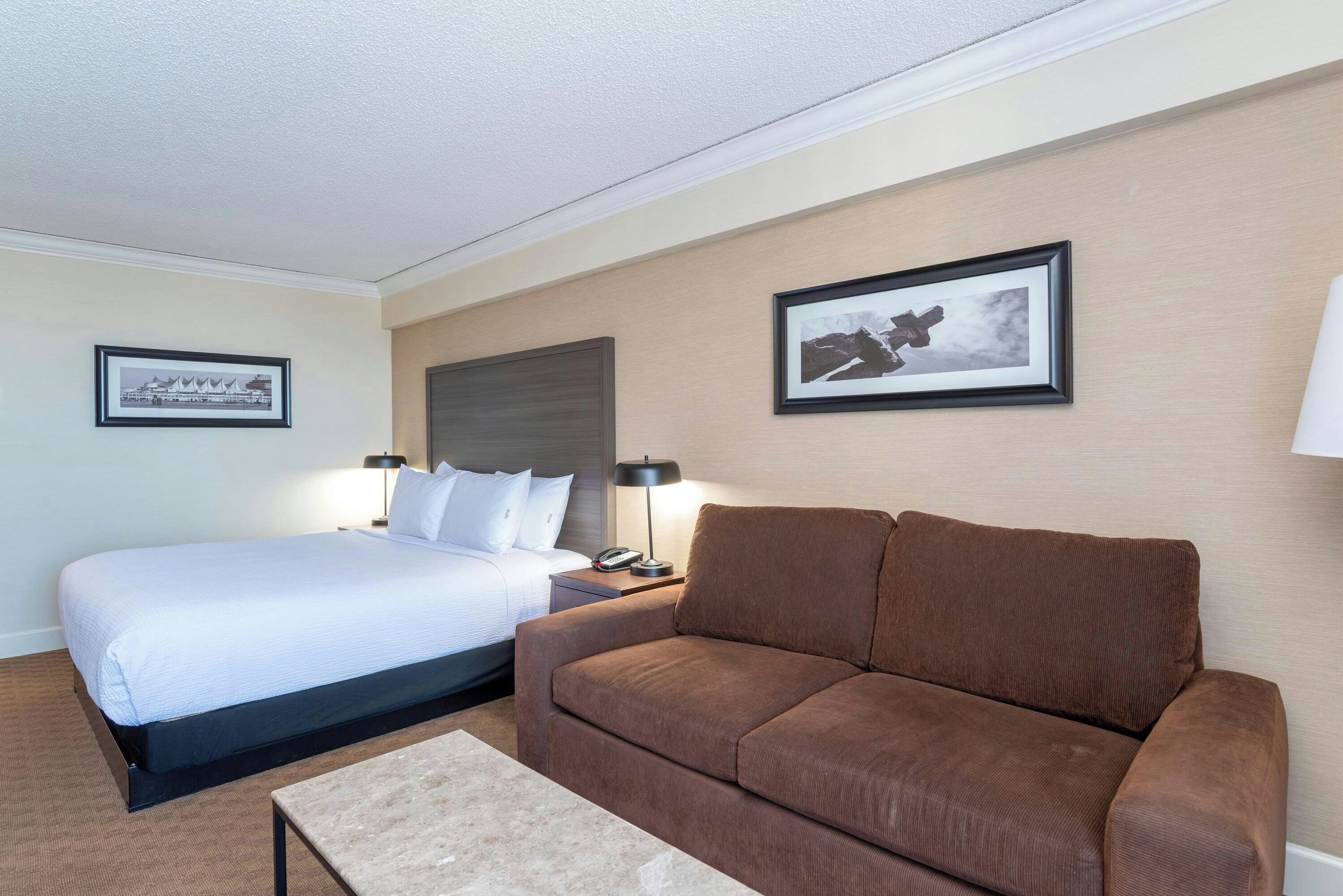 A hotel room with a bed, a brown sofa, a nightstand with a lamp, and two framed pictures on the wall.