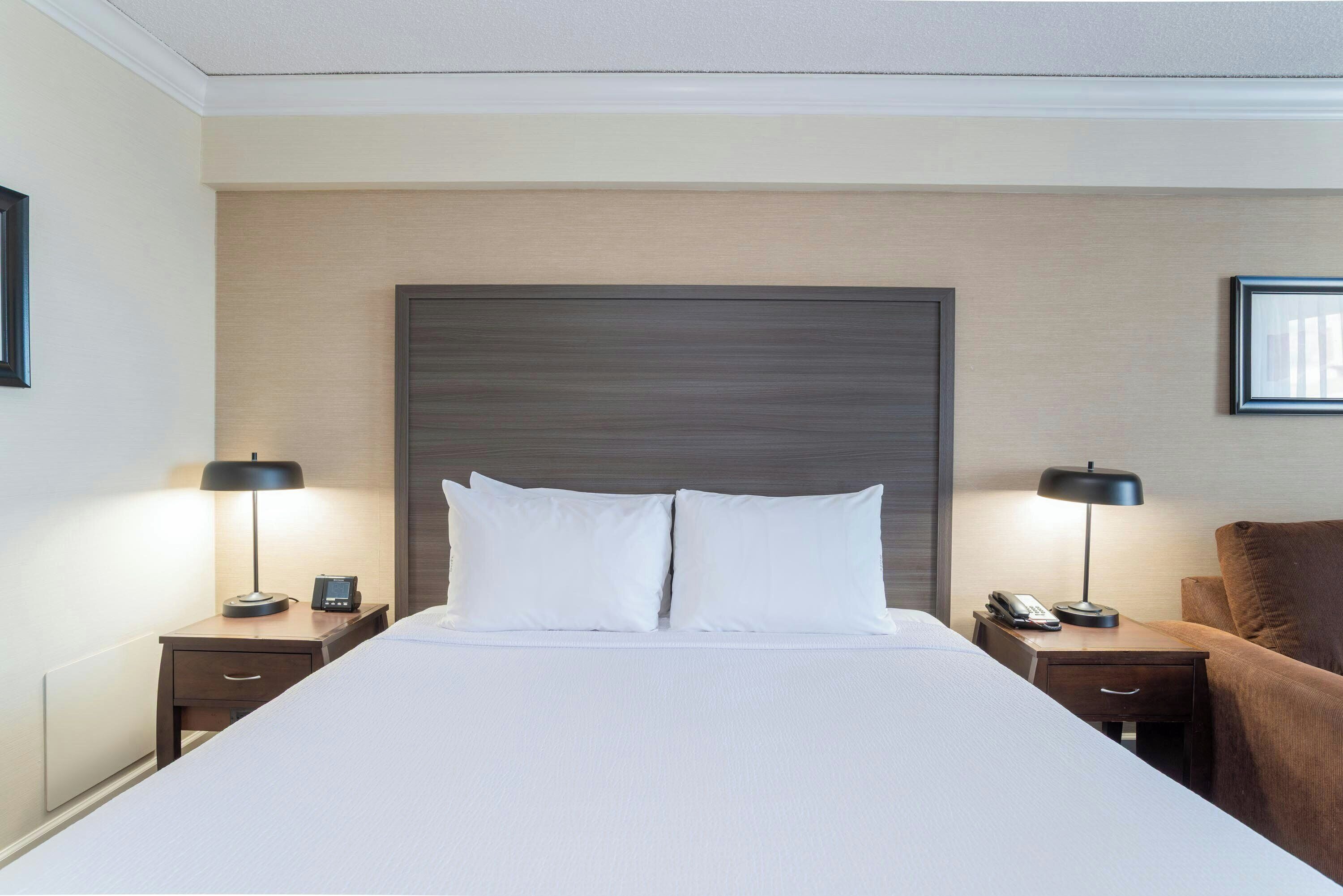 Quarto de hotel com uma cama grande, duas mesas de cabeceira, um criado-mudo, uma poltrona marrom e uma cabeceira.