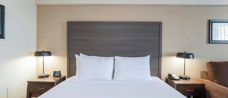 Quarto de hotel com uma cama grande, duas mesas de cabeceira, um criado-mudo, uma poltrona marrom e uma cabeceira.