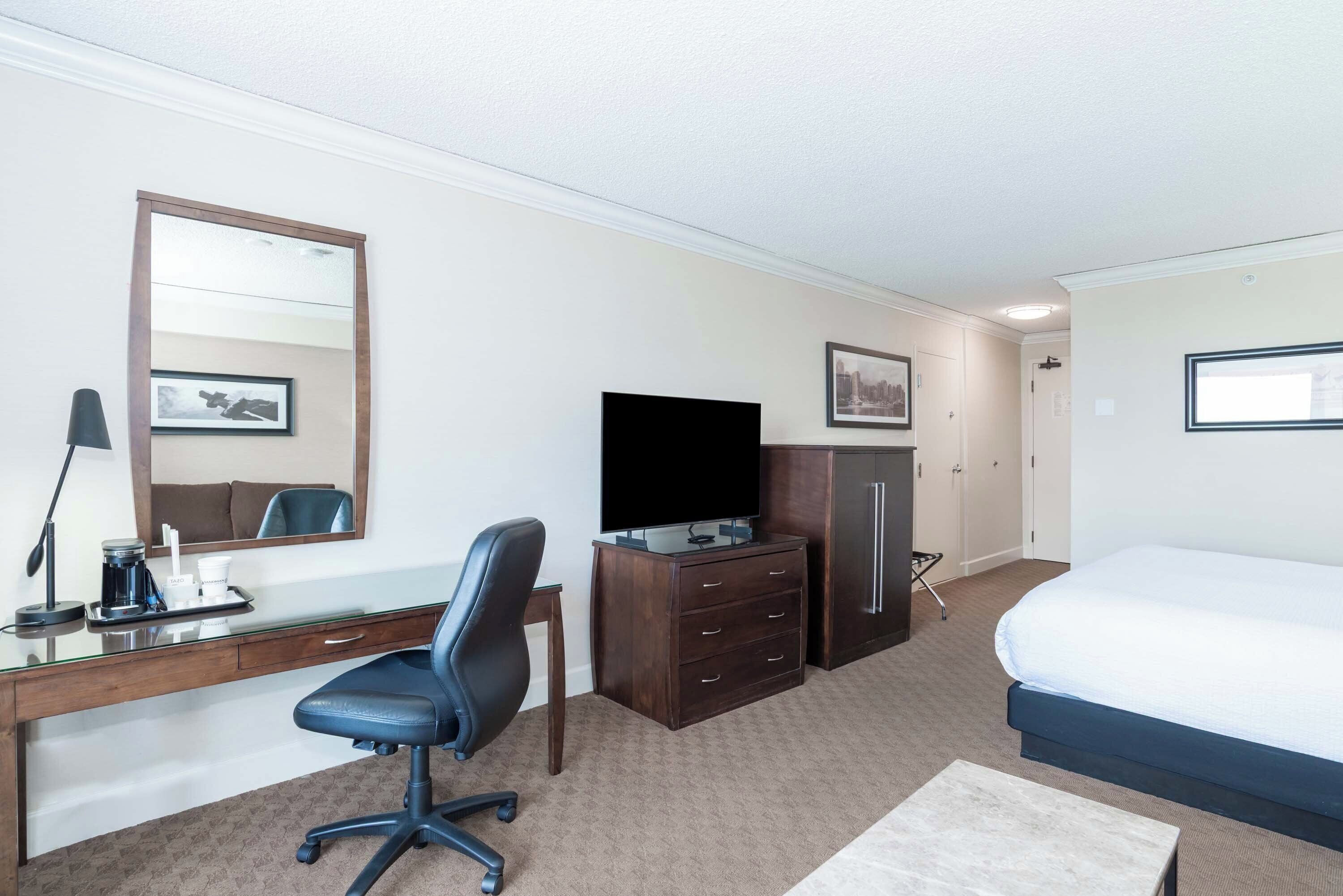 Una habitación de hotel con una cama, un escritorio con una silla, un televisor, un armario y un espejo.