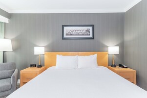 Una habitación de hotel con una cama grande, dos mesitas de noche con lámparas, un cuadro enmarcado en la pared y un sillón tapizado en gris.