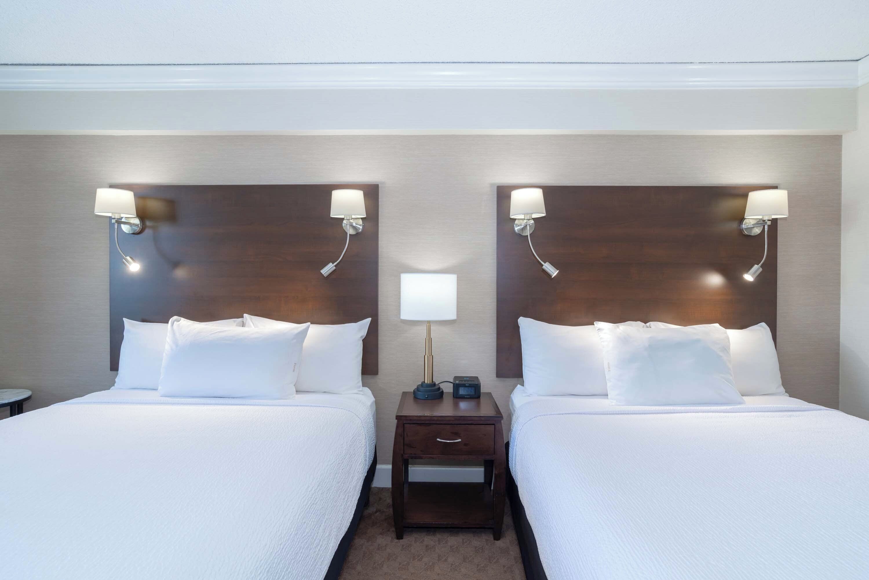 A hotel room with two beds, each with white linens and pillows, a nightstand with a lamp and a clock, and a wall-mounted light fixture.