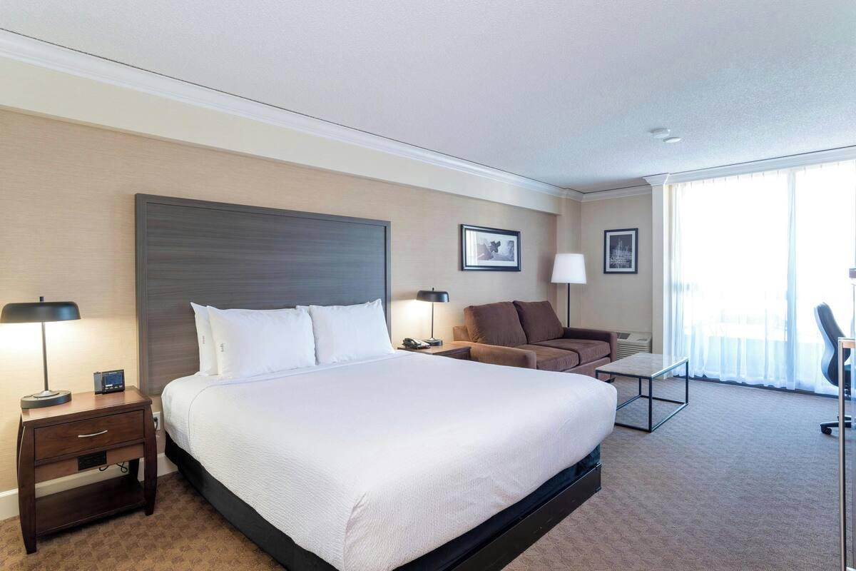 Une chambre d’hôtel avec un grand lit, une table de chevet, un bureau, une chaise et une fenêtre avec des rideaux.