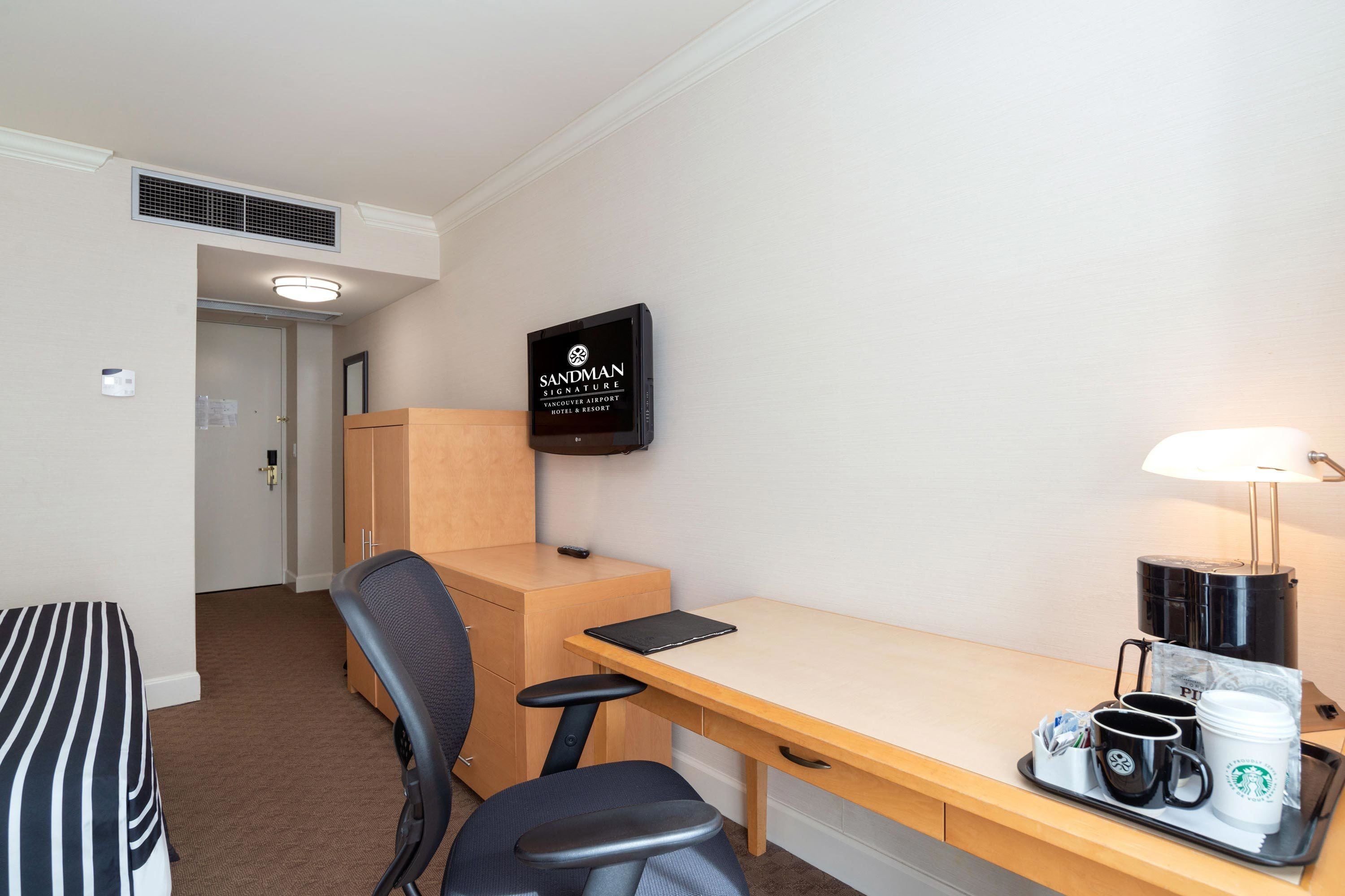 A hotel room with a bed, a desk, a chair, a TV, a lamp, and a tray with coffee cups and a Starbucks mug.