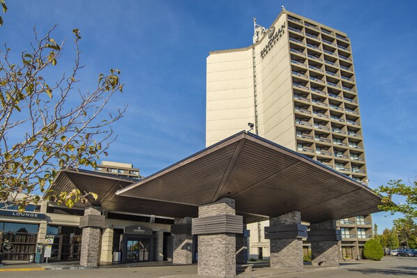 A hotel entrance with a covered lobby area, a stone facade, and a tall building in the background. - Sandman Signature Vancouver Airport Hotel & Resort (Richmond)