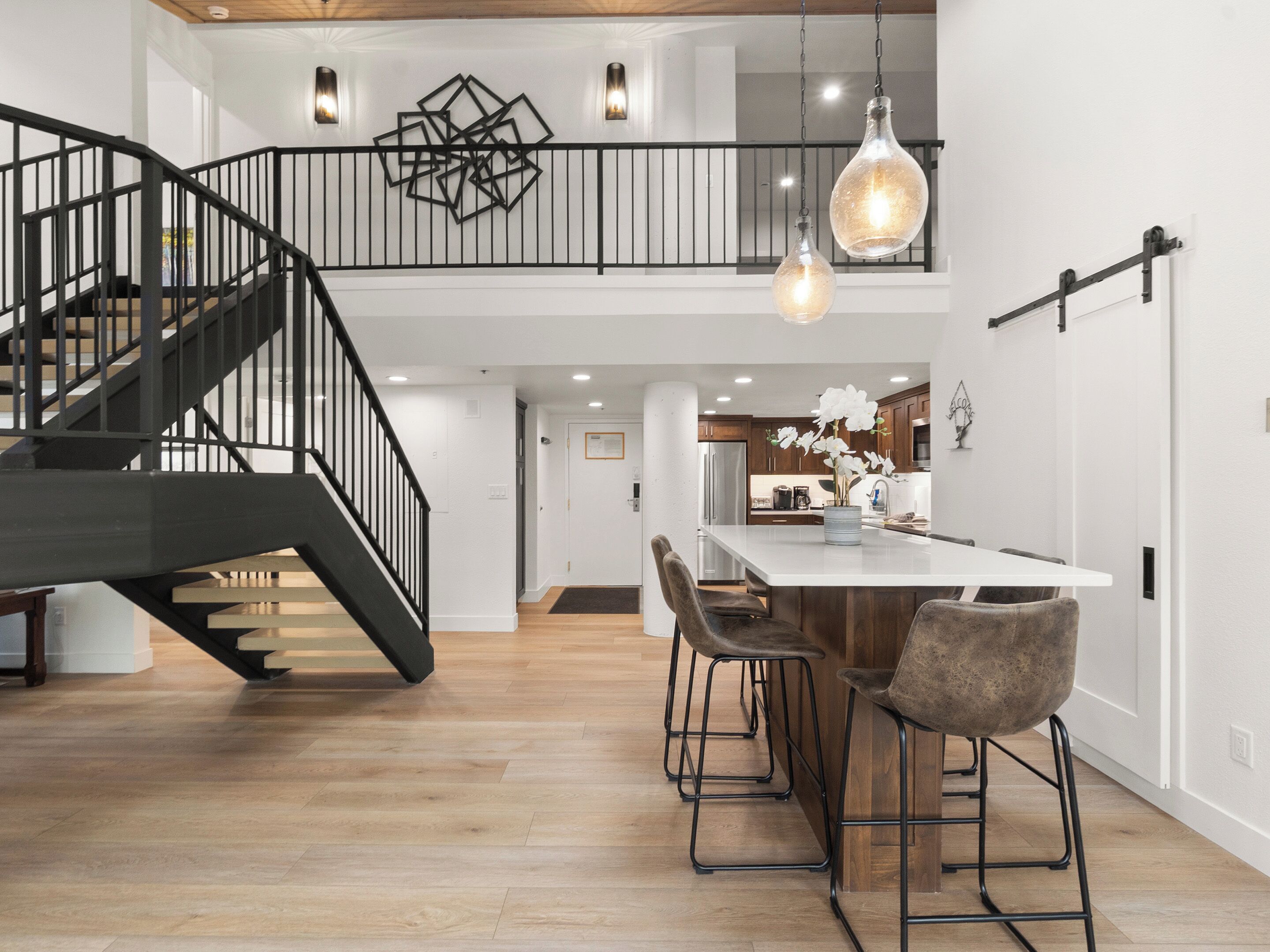 A spacious kitchen with a central island, a staircase with black railings, and pendant lighting.