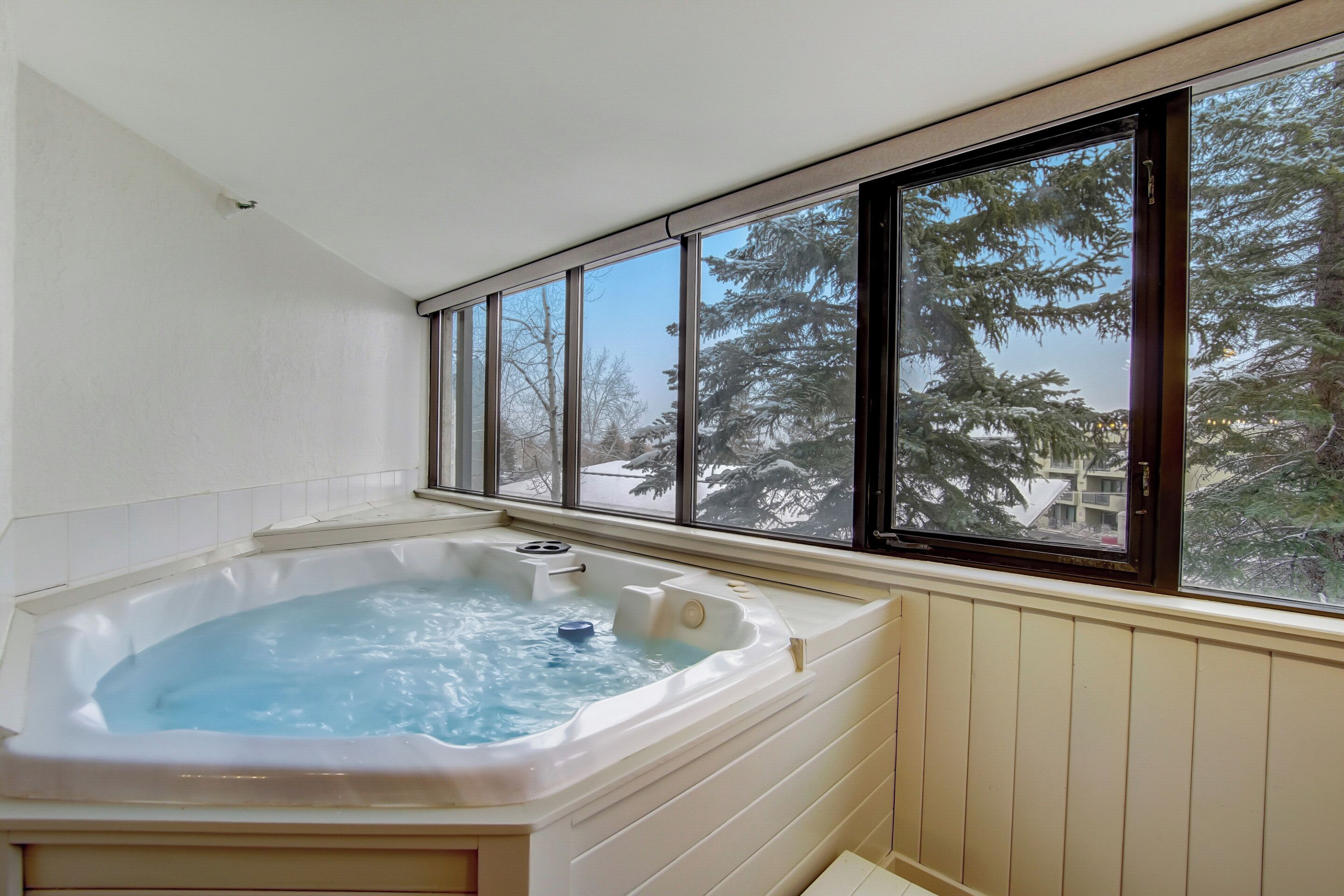 A hot tub with a view of trees and buildings through large windows.