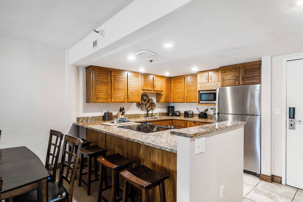 Cocina moderna con armarios de madera, refrigerador de acero inoxidable, encimeras de granito y una isla central con taburetes de bar.