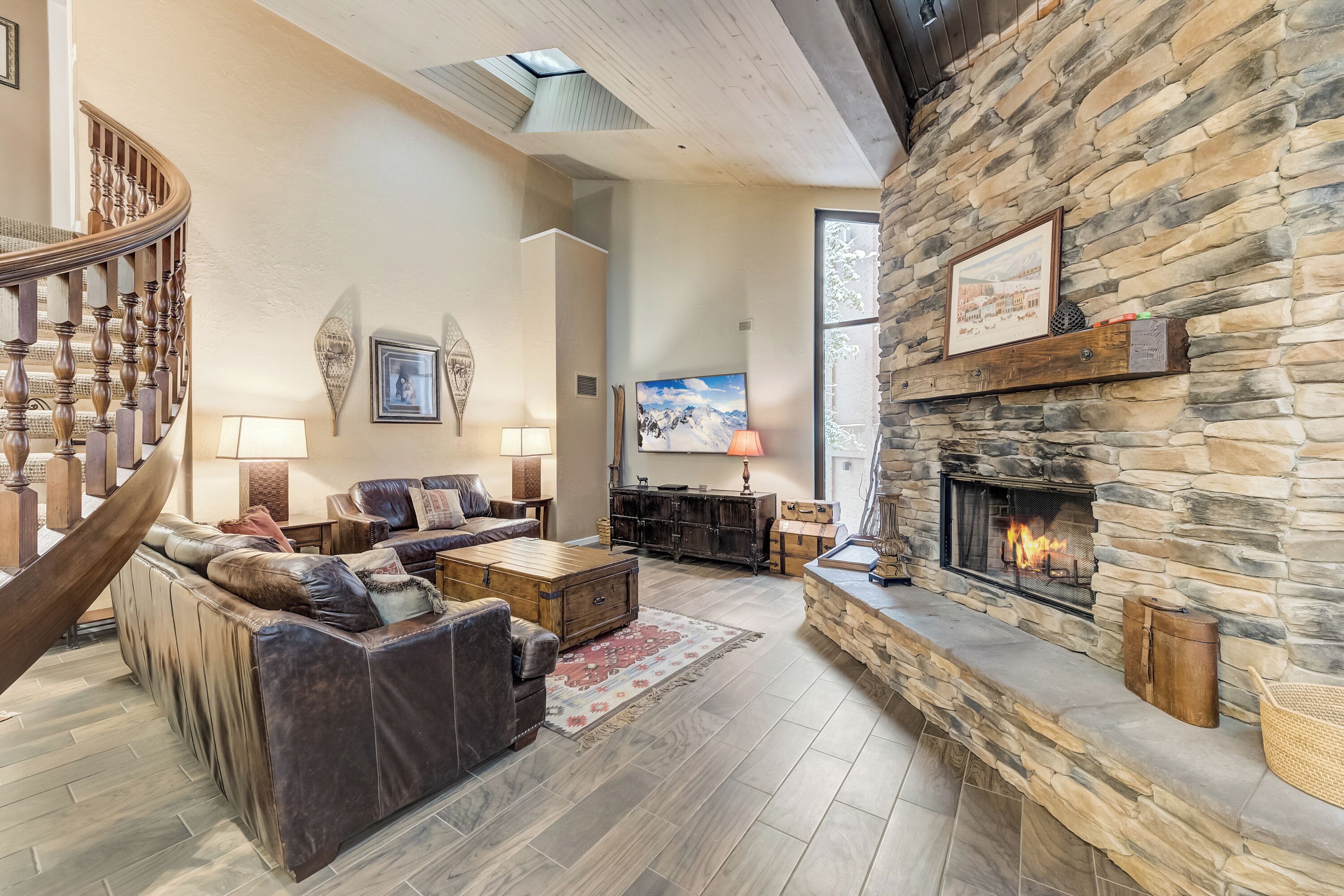 A spacious living room with a stone fireplace, leather sofa, and a wooden staircase.