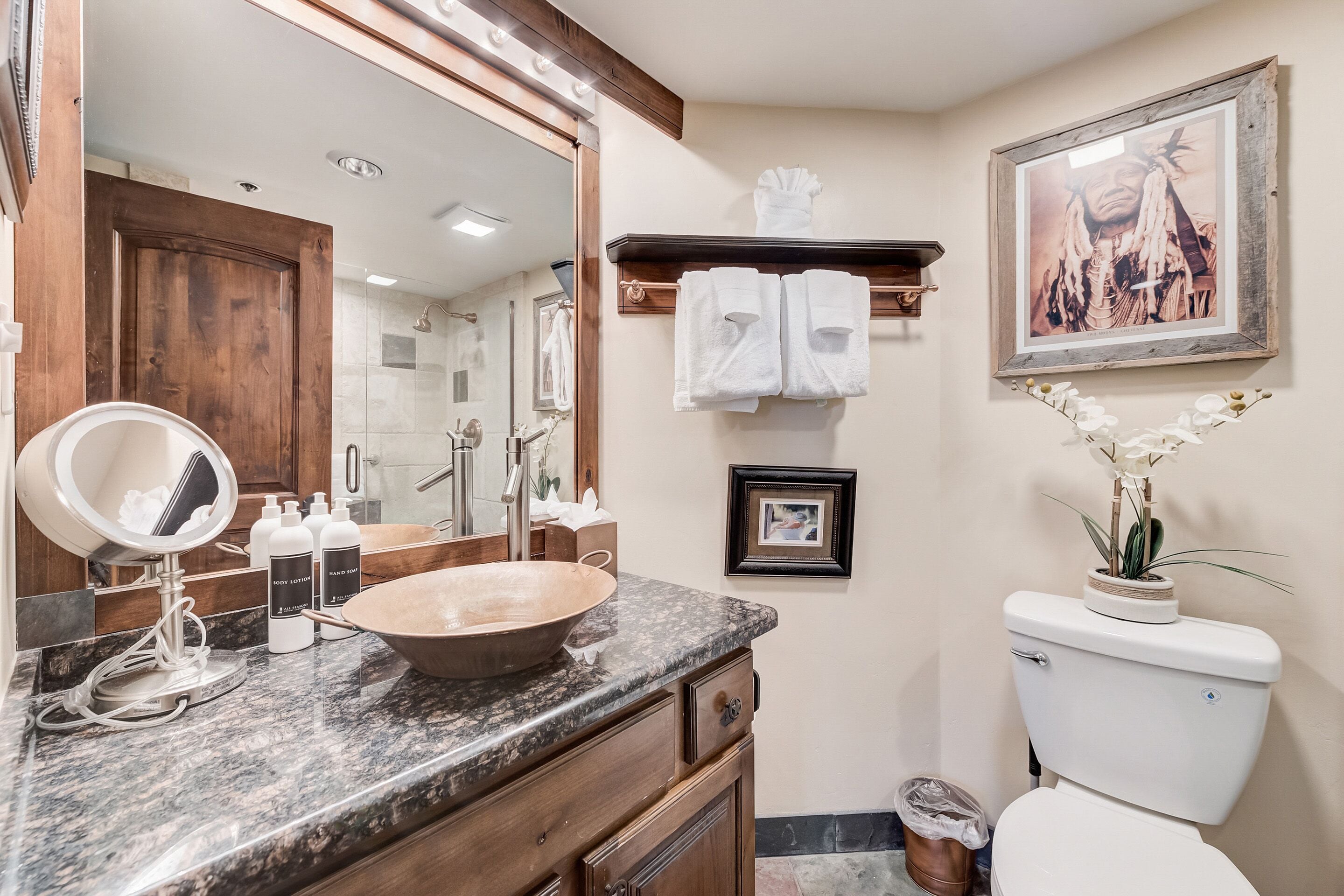 Une salle de bain avec un meuble-lavabo en bois, un grand miroir, un lavabo sur colonne et des toilettes. Des serviettes sont suspendues sur un porte-serviettes et un tableau encadré est accroché au mur.