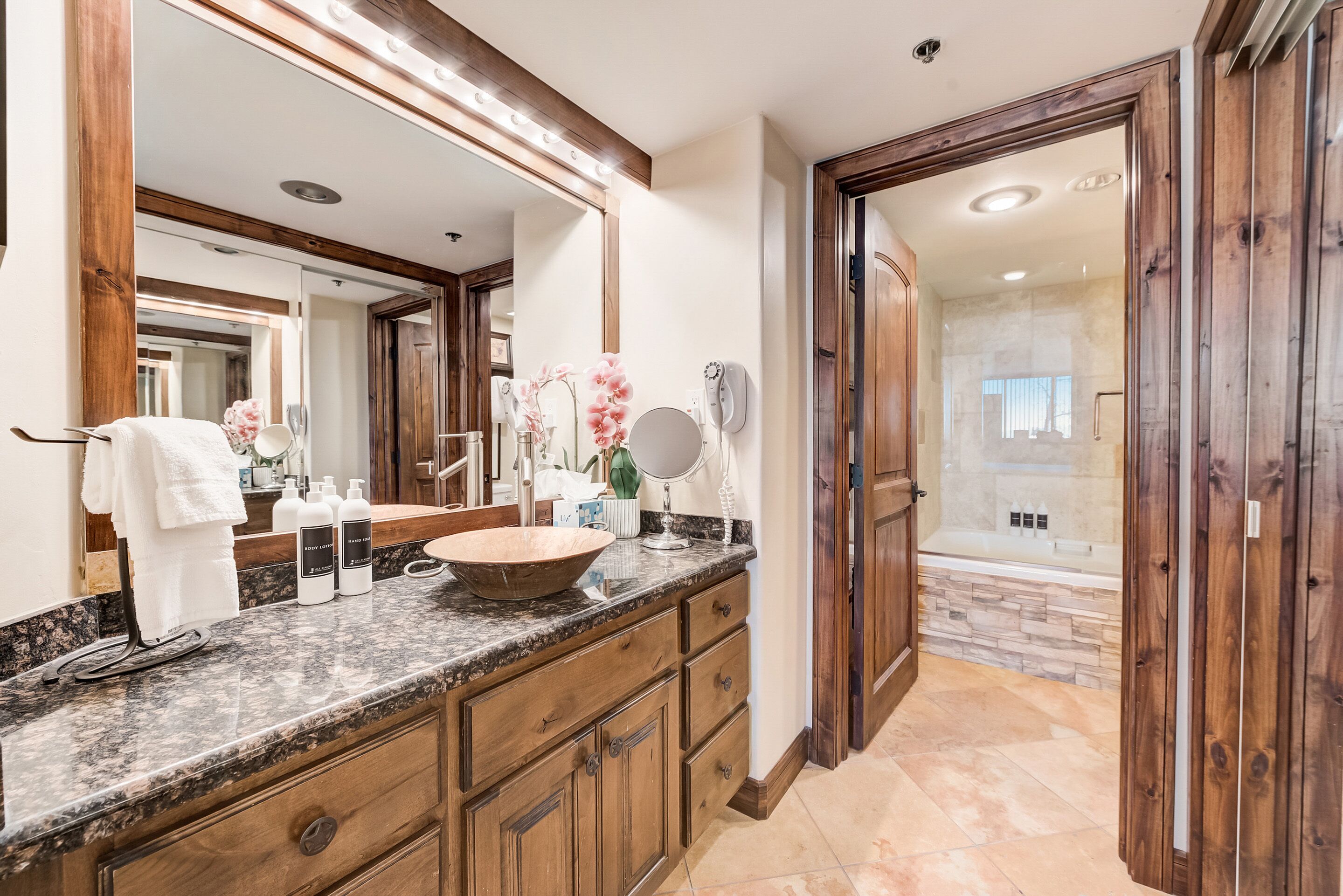 A bathroom with a large mirror, wooden cabinets, a marble countertop, and a shower area visible through an open door.