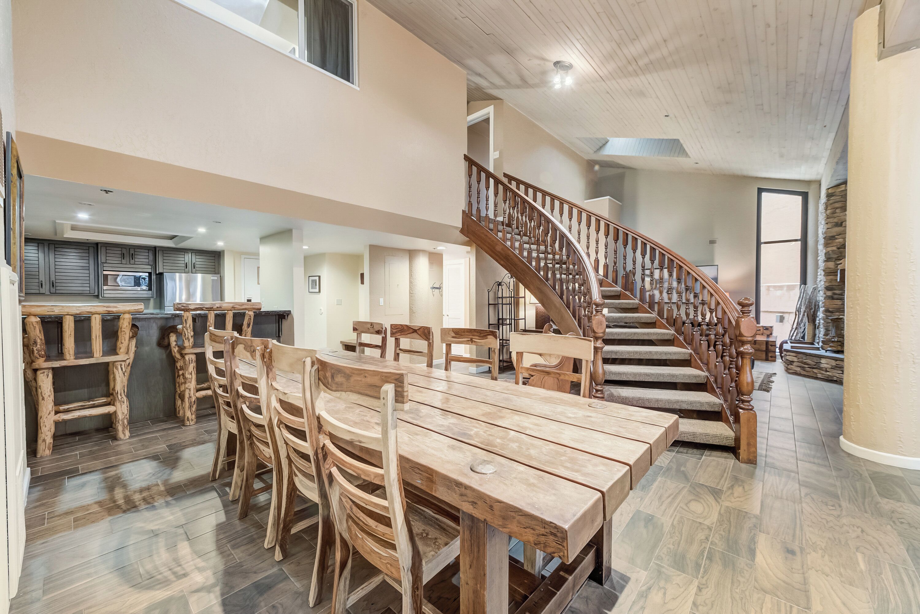 A spacious dining area with a long wooden table and chairs, a wooden staircase, and a kitchen in the background.