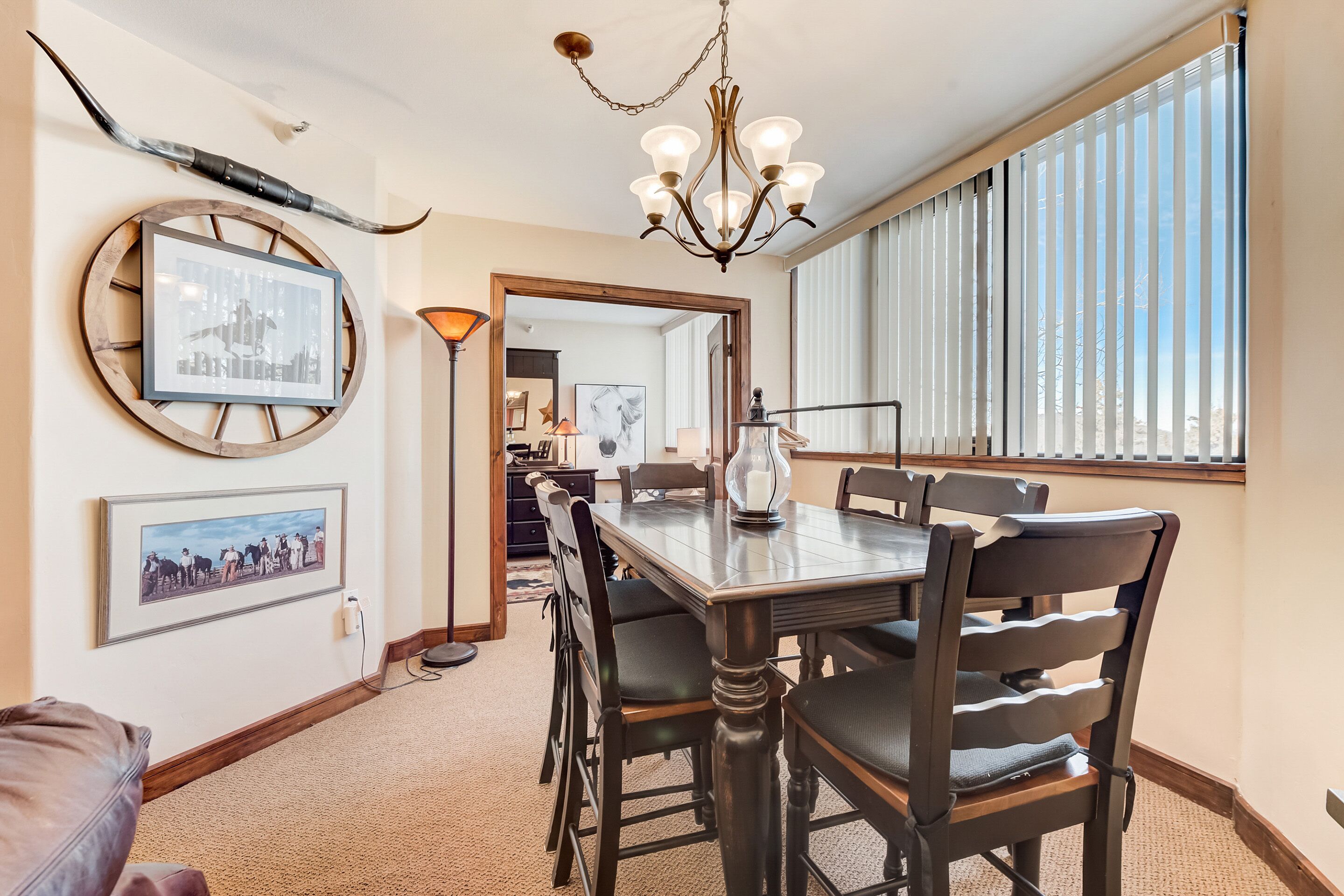 A dining area with a wooden table and chairs, a chandelier, framed pictures on the wall, and a large window with blinds.