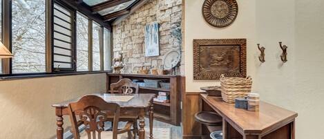A dining area with a wooden table, chairs, a patterned rug, a stone wall, and a window with a view of trees.
