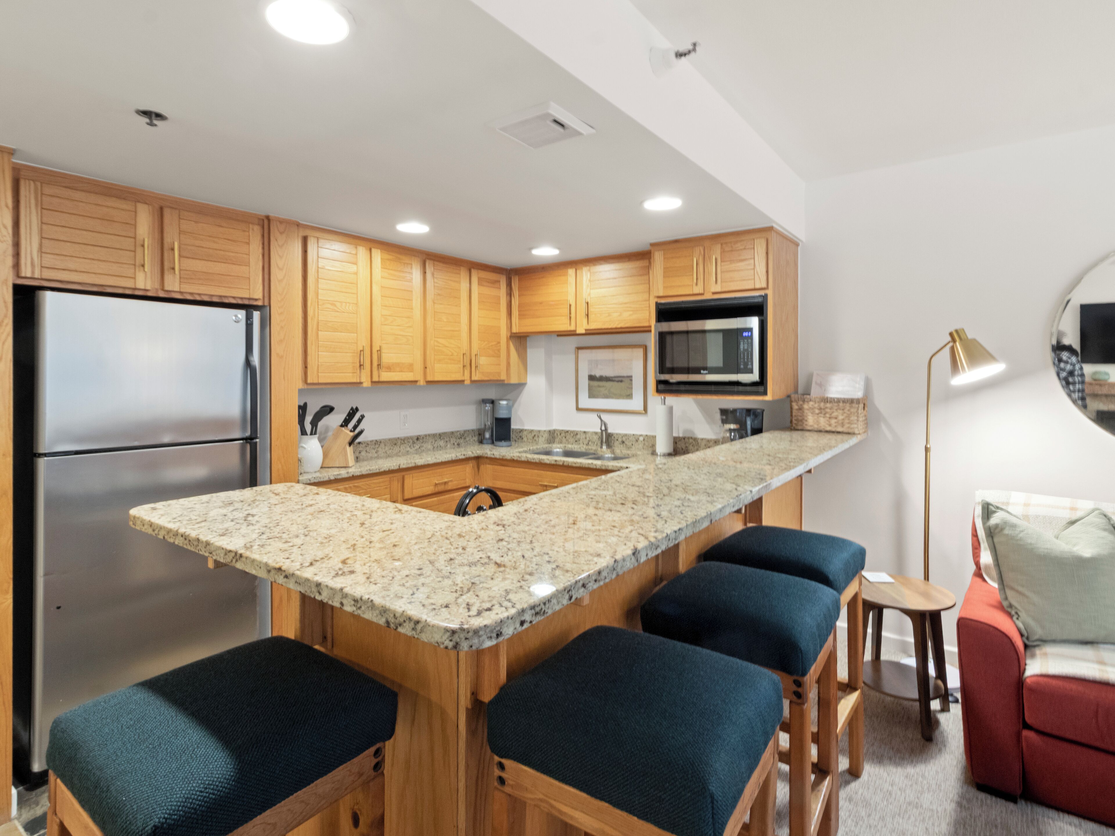 A modern kitchen with wooden cabinets, a stainless steel refrigerator, a granite countertop, and a bar seating area with blue cushioned stools.