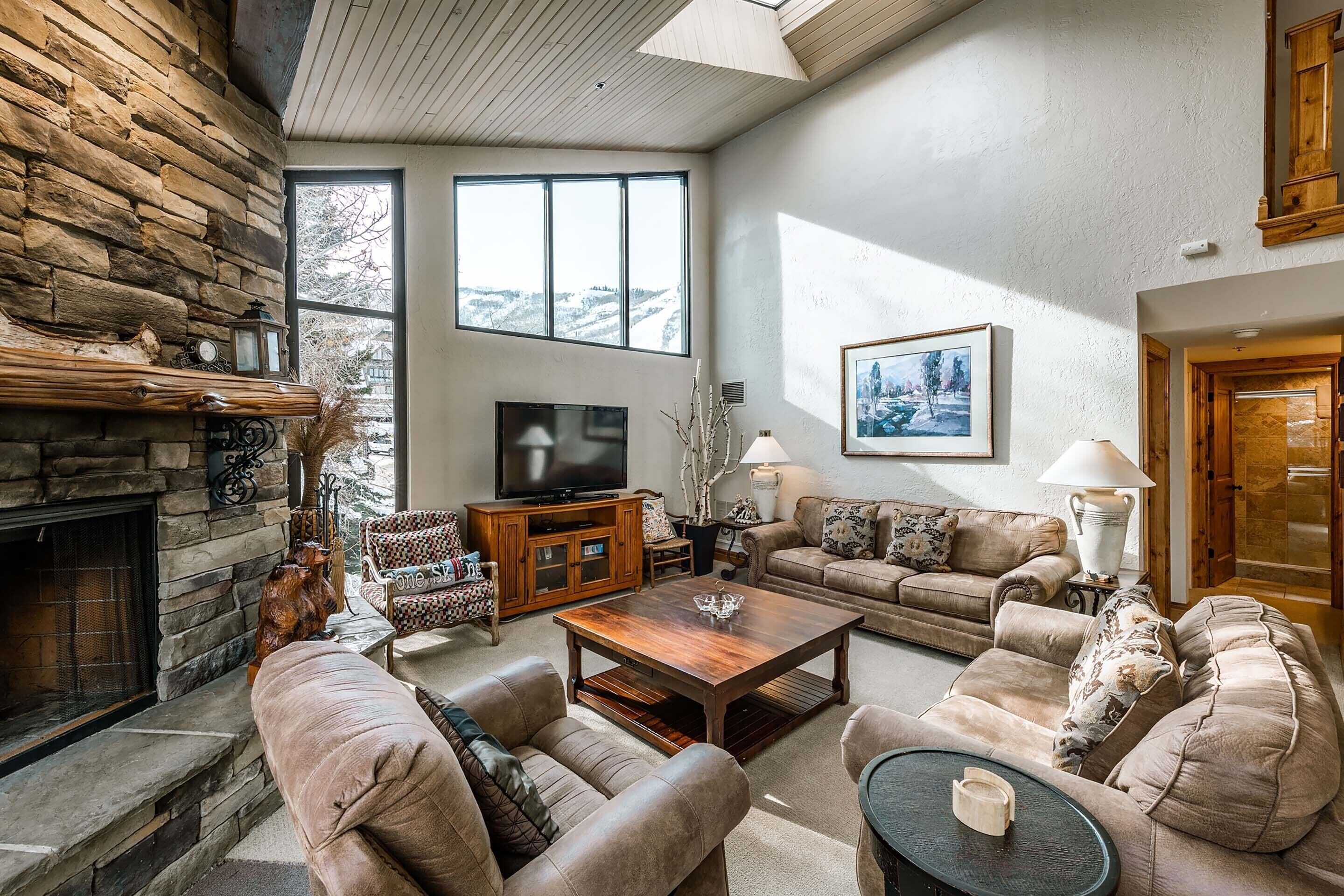 A cozy living room with a stone fireplace, comfortable seating, a flat-screen TV, and large windows offering a view of the outdoors.