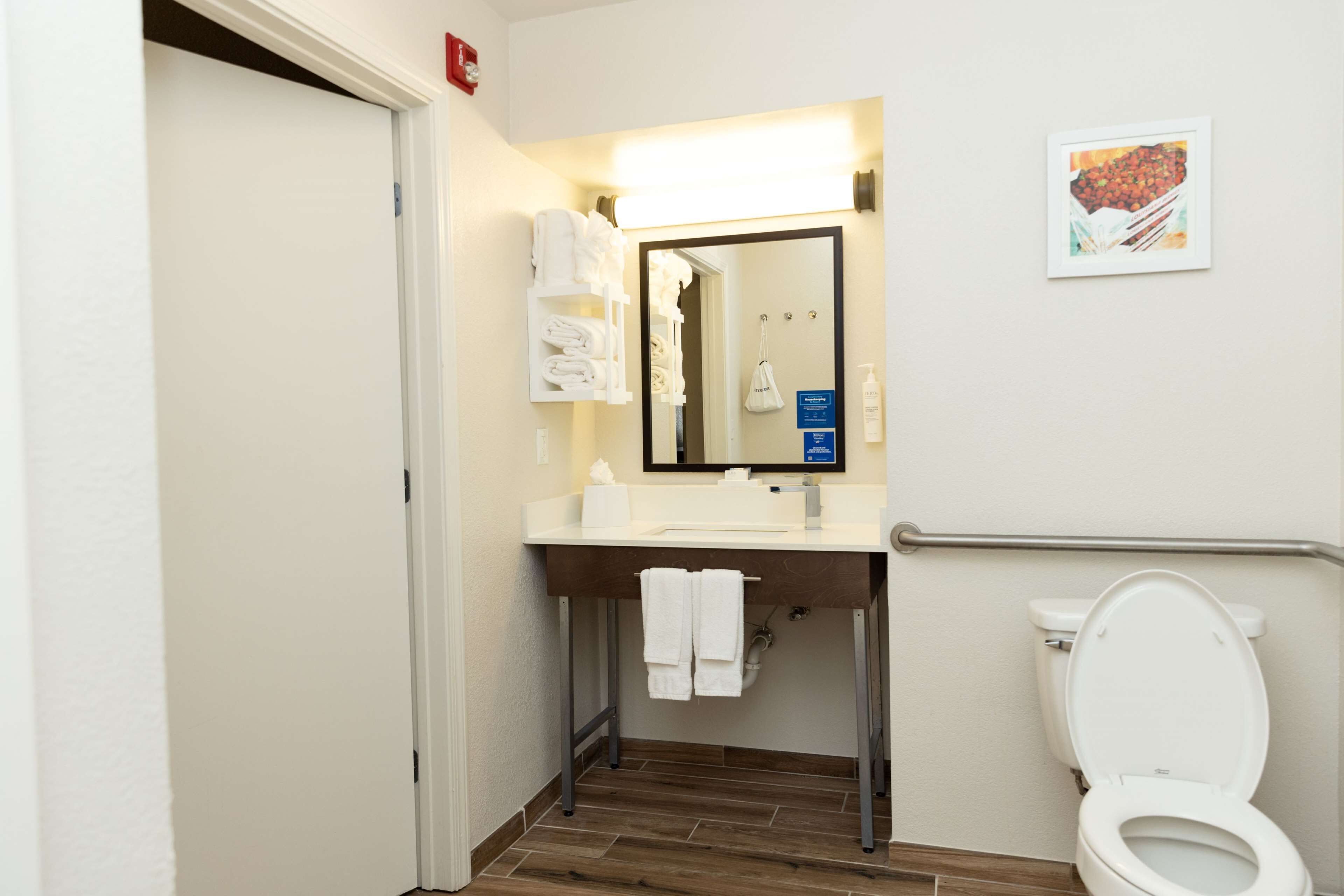 A small, clean bathroom with a white sink, mirror, and toilet. There are towels hanging on the wall and a framed picture above the toilet.