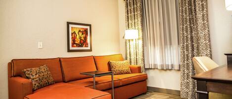 Ein Hotelzimmer mit Ecksofa, einem kleinen Tisch, einer Lampe, gemusterten Vorhängen und einem gerahmten Bild an der Wand.