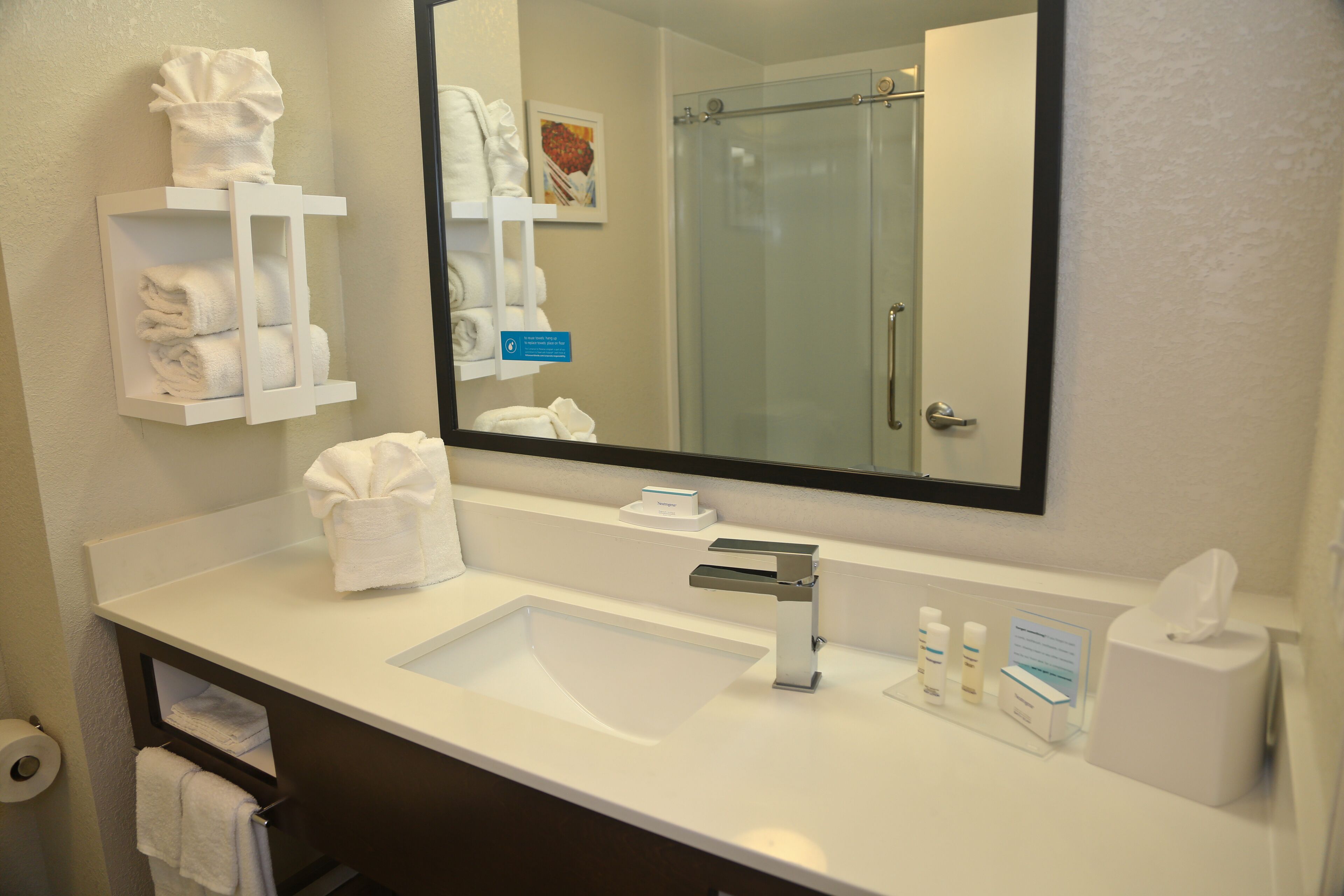 A hotel bathroom with a white sink, a mirror, and shelves holding towels.