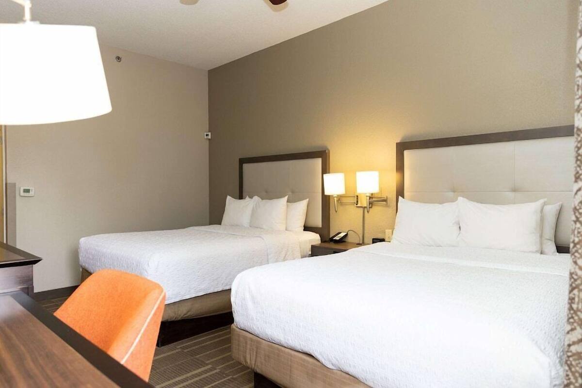 A hotel room with two beds, a desk, an orange chair, and bedside lamps.