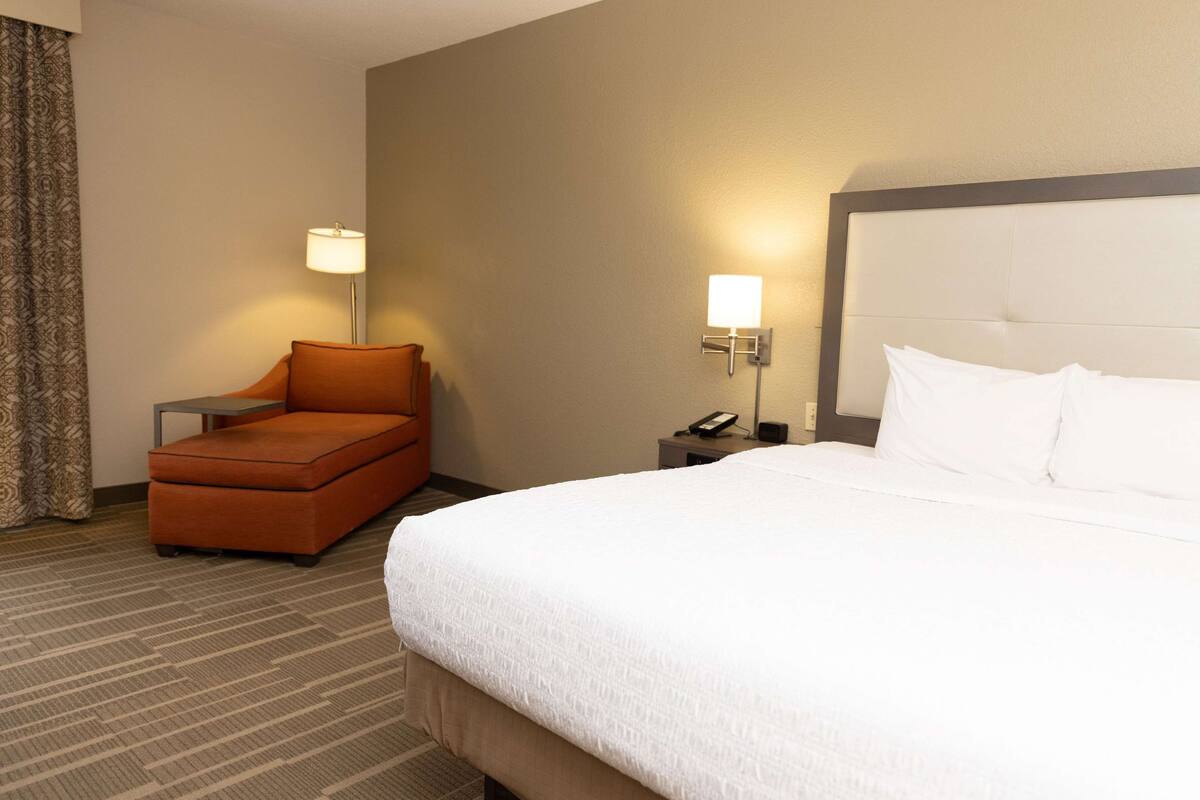 A hotel room with a large bed, a small orange armchair, a bedside table with a lamp, and a phone.
