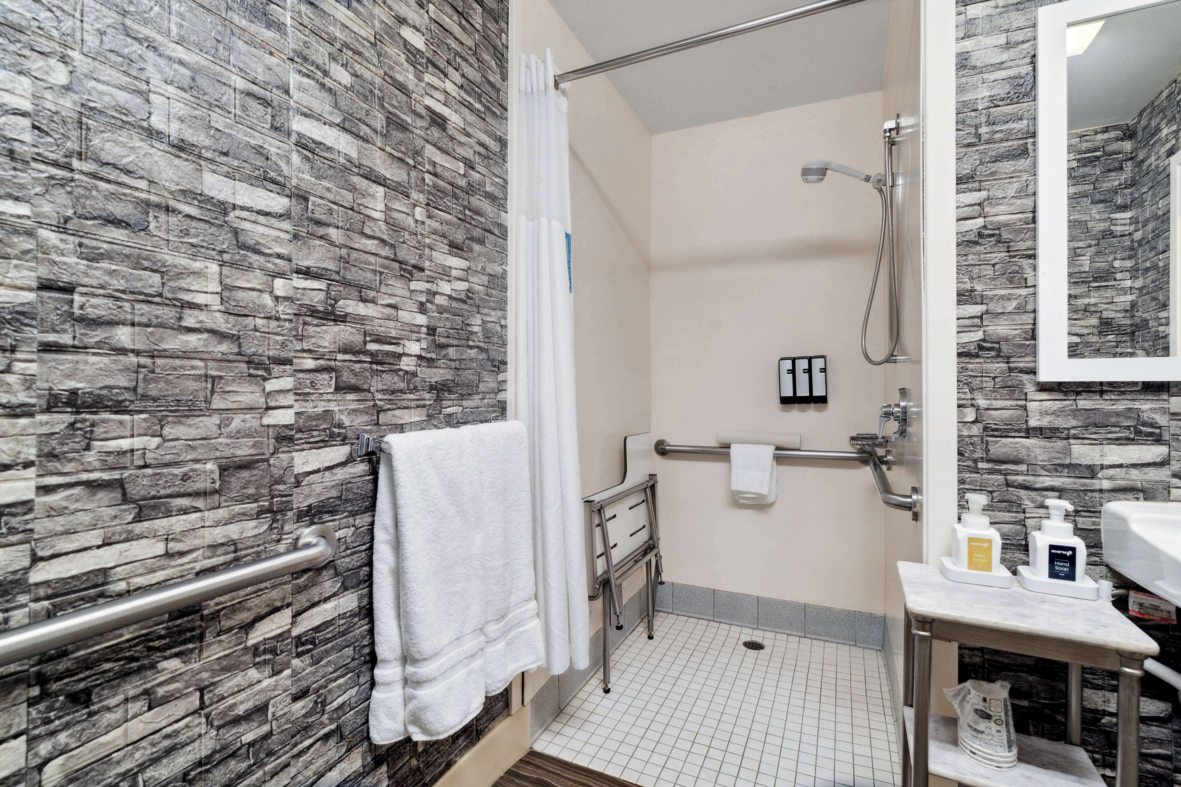 Un baño con pared de piedra, ducha, barra de apoyo, toallero y lavabo con artículos de aseo.