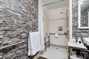 Un baño con pared de piedra, ducha, barra de apoyo, toallero y lavabo con artículos de aseo.