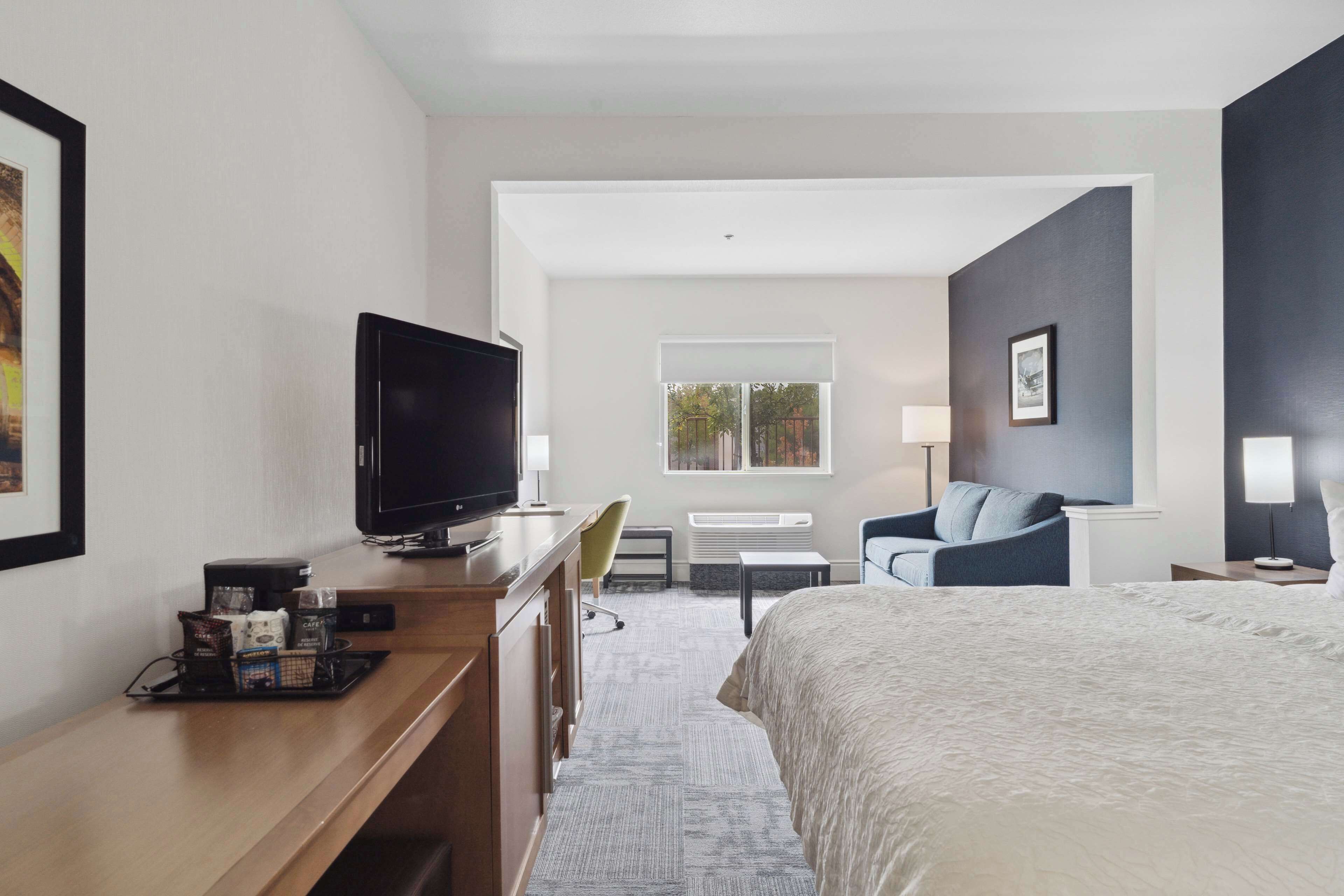 A hotel room with a bed, a desk with a TV, a chair, a lamp, and a window.