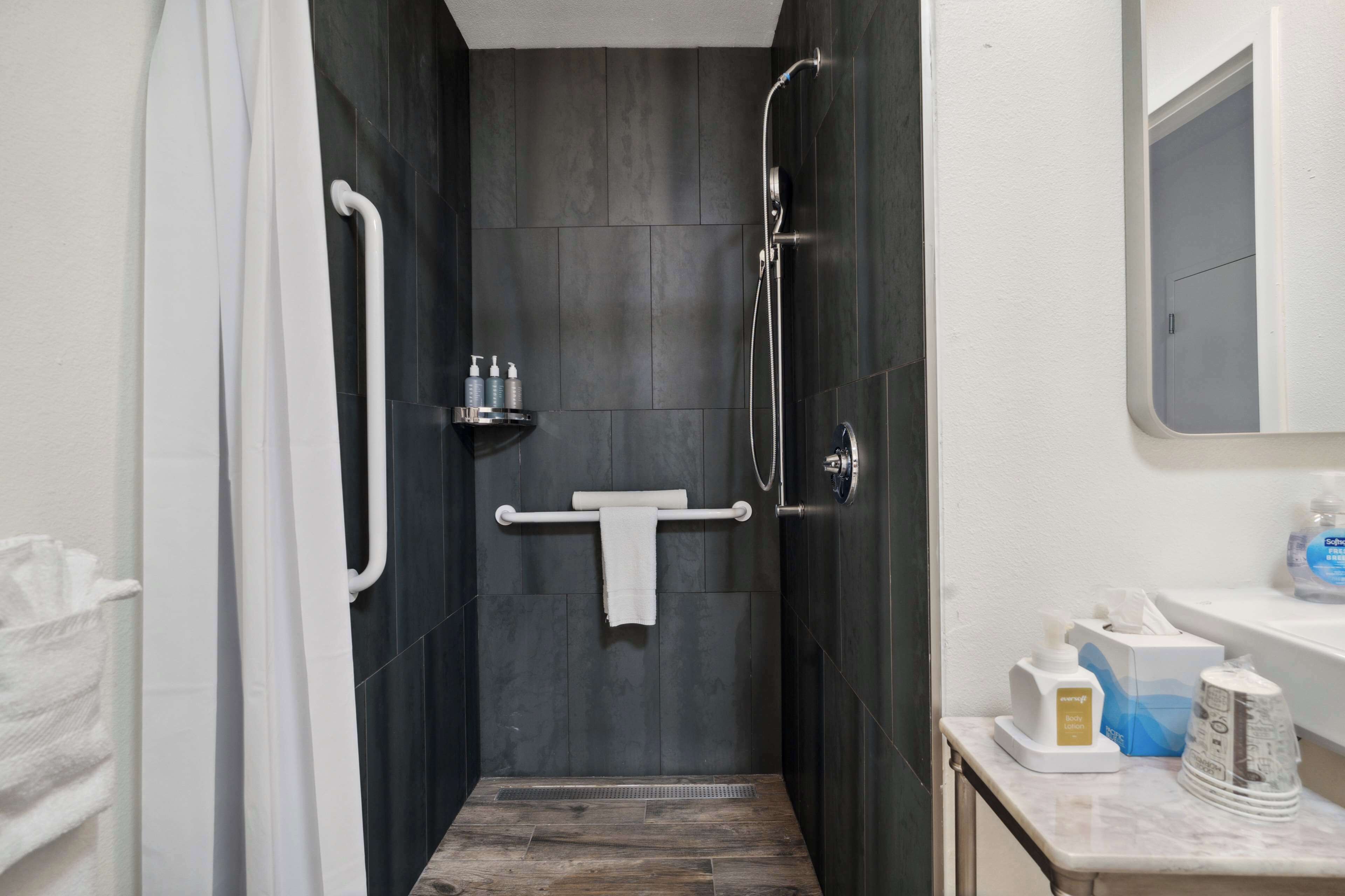 Une salle de bain moderne avec une douche à l’italienne, des murs recouverts de carreaux foncés, un porte-serviettes blanc et un plan de toilette avec des articles de toilette.