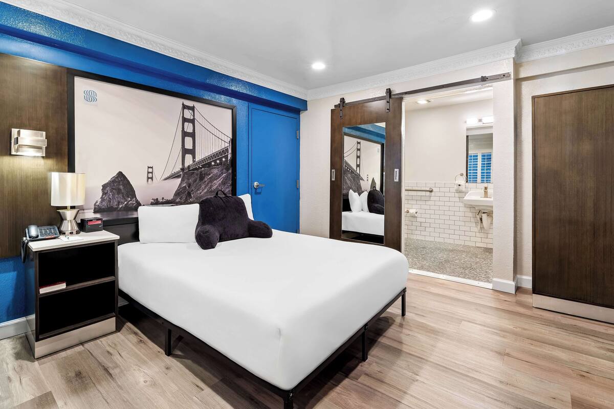 Ein modernes Hotelzimmer mit einem großen Bett, einem Nachttisch mit Lampe, einem Schreibtisch mit Telefon und Kaffeemaschine sowie einem großen, gerahmten Bild der Golden Gate Bridge.