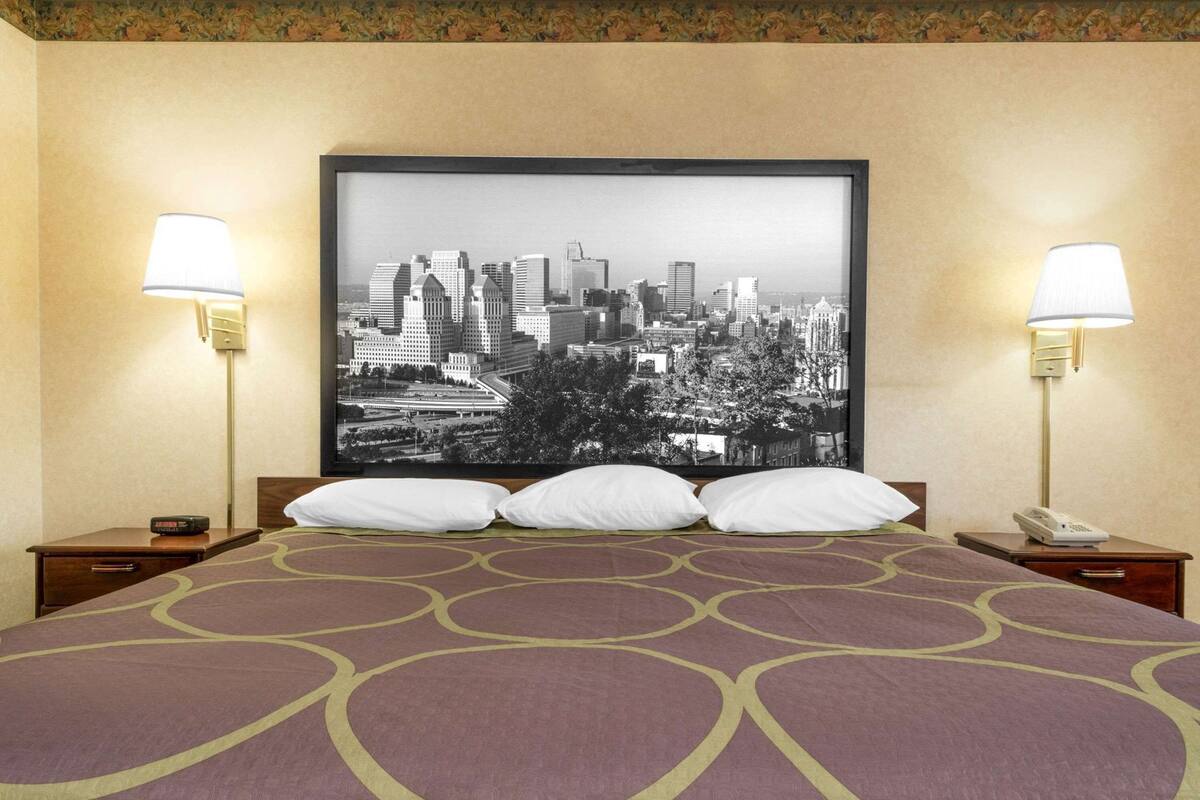 A hotel room with a large bed, two bedside lamps, a framed cityscape picture, and a nightstand with a phone.