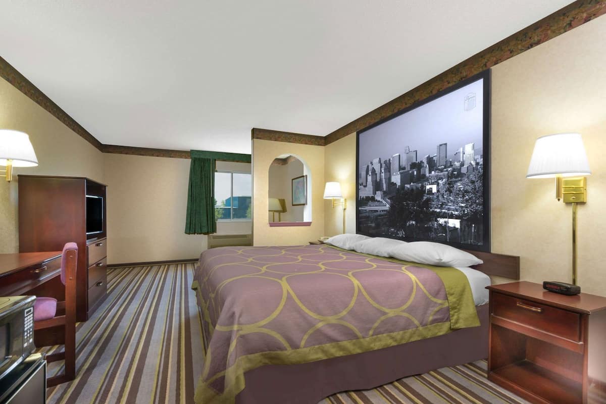 A hotel room with a bed, a desk, a chair, a TV, a lamp, and a cityscape painting on the wall.