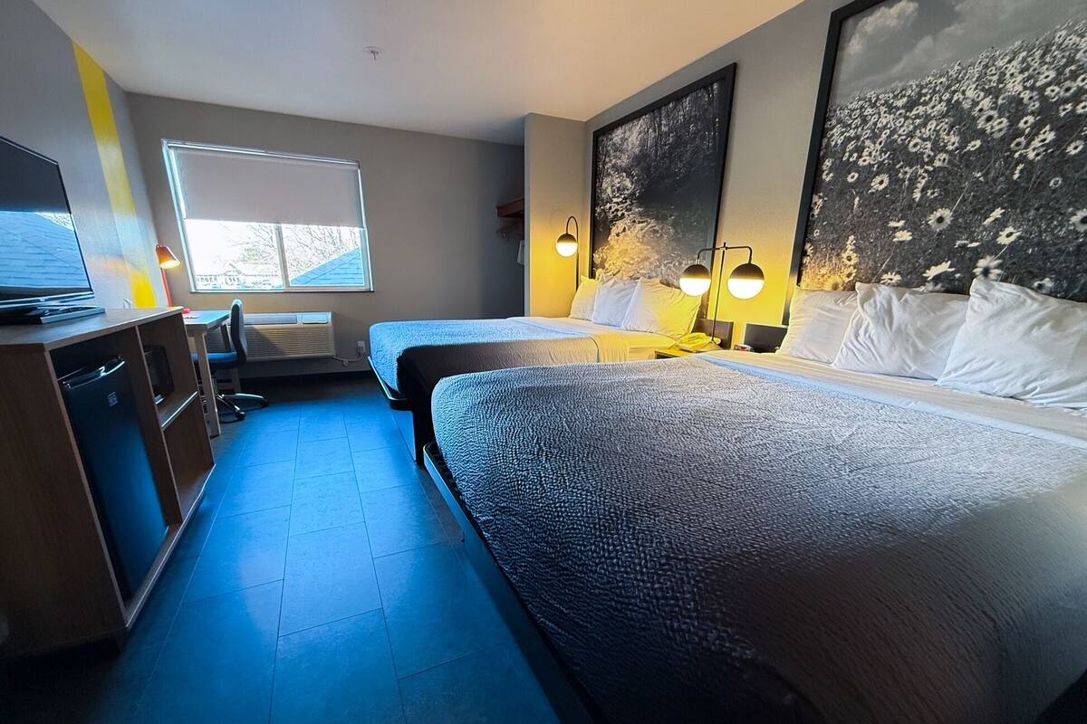 Ein Hotelzimmer mit zwei Betten, einem Schreibtisch, einem Stuhl, einem Fernseher und einem großen Fenster.
