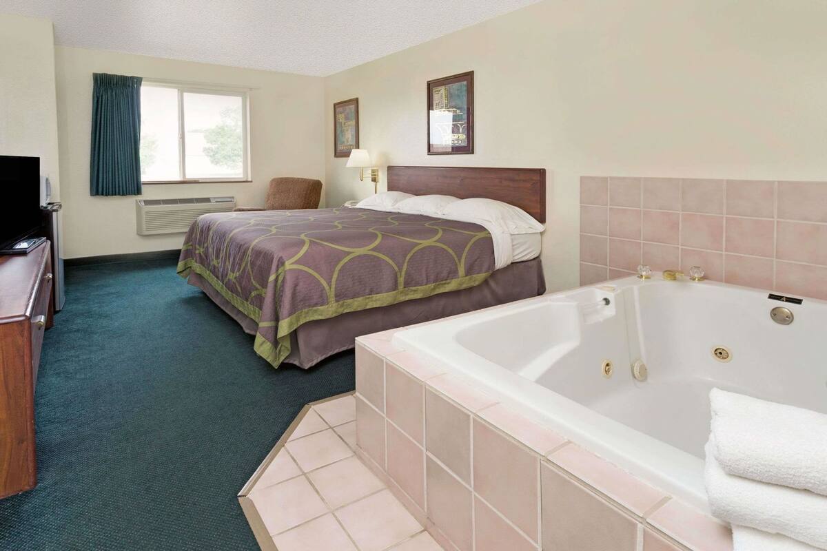 Ein Hotelzimmer mit Bett, Schreibtisch, Stuhl, Fenster, Wandlampe, einem gerahmten Bild und einer Badewanne.