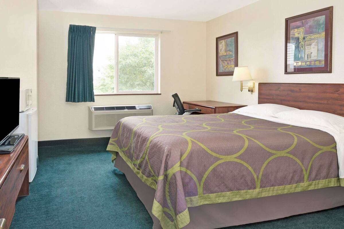 Ein Hotelzimmer mit einem Bett, einem Schreibtisch, einem Stuhl, einem Fernseher, einem Fenster mit Vorhängen, einer Klimaanlage und zwei gerahmten Bildern an der Wand.