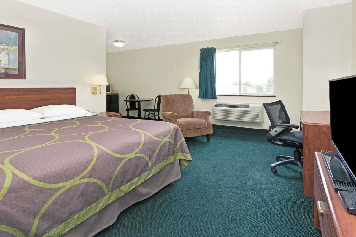 Ein Hotelzimmer mit einem großen Bett, einem Schreibtisch mit Computer, einem Sessel, einem kleinen Tisch und einem Fenster mit Vorhängen.
