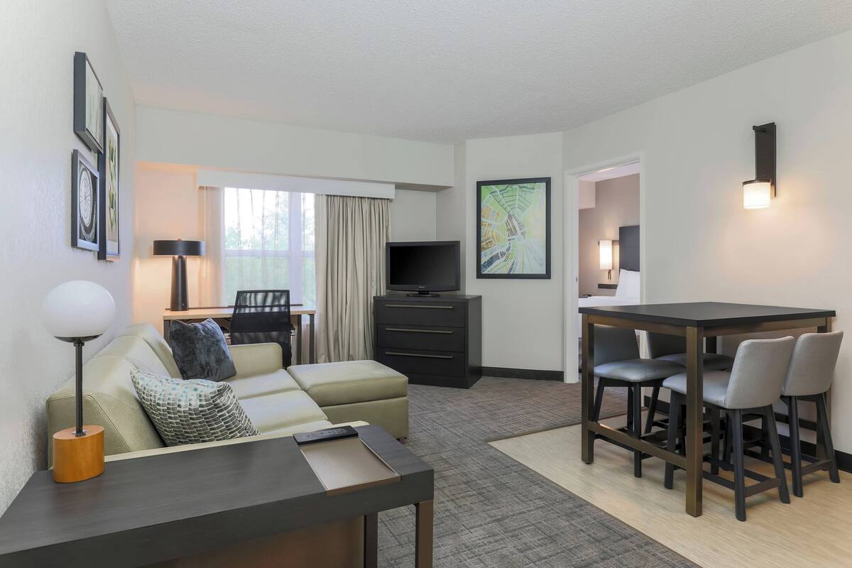 A modern hotel room with a sofa, a dining table with chairs, a TV on a stand, and a map on the wall.
